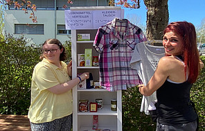 Auf dem Schulhof mit dem Schulgebäude im Hintergrund steht ein weißes Regal mit sechs Etagen. Darin befinden sich verschiedene Lebensmittel (Tee in Geschenkverpackungen, Suppen in Konservendosen, Schokoladentafeln und andere), die gratis verteilt werden.