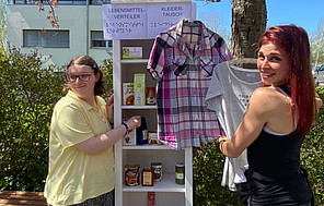Die Aufklärer*innen Auf dem Schulhof mit dem Schulgebäude im Hintergrund steht ein weißes Regal mit sechs Etagen. Darin befinden sich verschiedene Lebensmittel (Tee in Geschenkverpackungen, Suppen in Konservendosen, Schokoladentafeln und andere), die gratis verteilt werden.